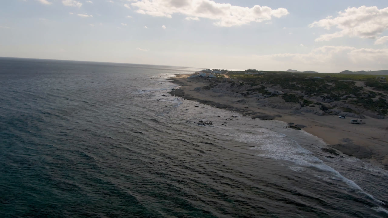 Drone perspective at Shipwrecks Beach in Los Cabos, showing rippled blue water, white surf bands, a sandy cove, rocky terrain, and no visible people.