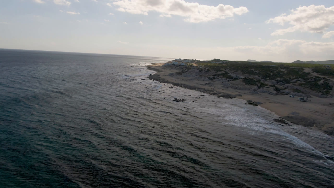 Wide aerial coastline with ocean foreground, beach midground, and low hills beyond.