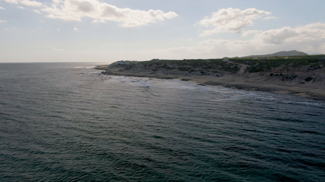 Opening aerial frame over Shipwrecks Beach in Los Cabos, with dark ocean in the foreground, surf along the sandy shore, low cliffs, distant hills, clouded sky, and no visible people.