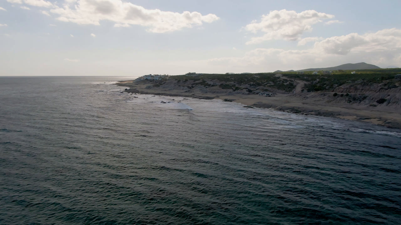 Aerial view at Shipwrecks Beach in Los Cabos showing more of the shoreline curve, white surf bands, rocky patches near the water, low cliffs, distant hills, cloudy sky, and no visible people.