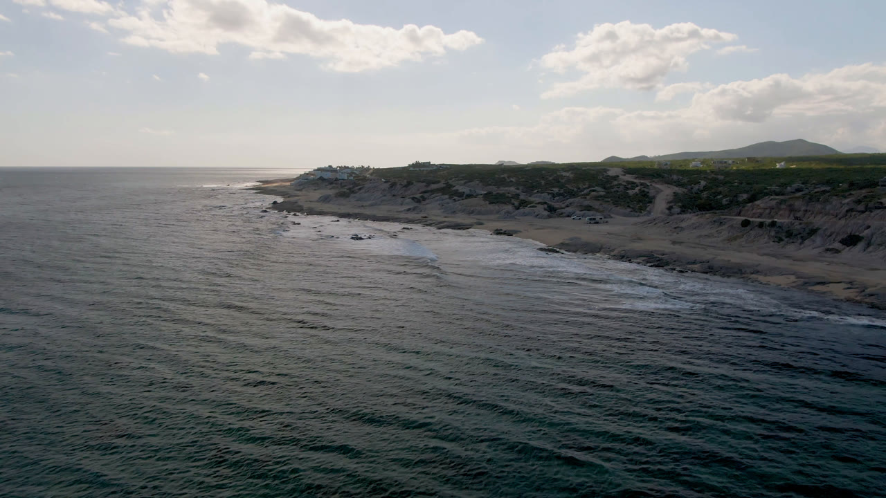 Ocean and rocky beach with cliffs, green land, hills, and bright clouds.