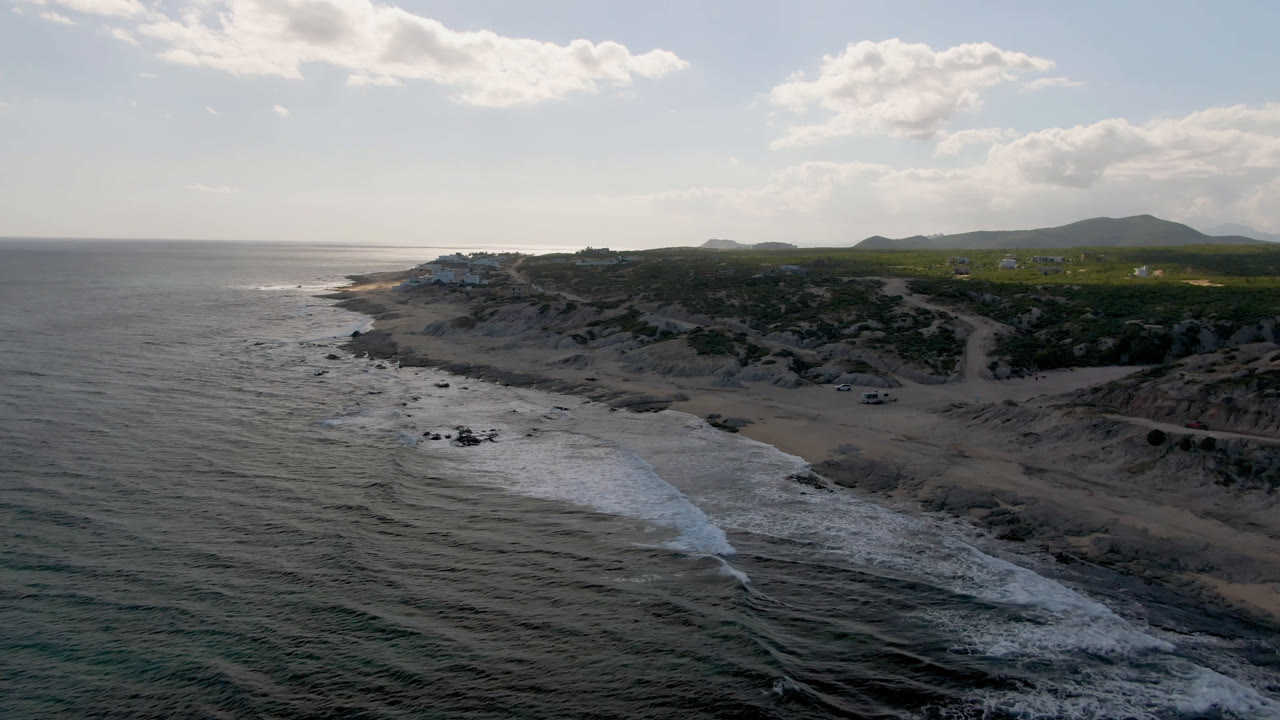 Aerial view of Shipwrecks Beach in Los Cabos with surf entering from the lower left, sandy dunes and scrub across the shore, distant hills on the horizon, and no visible people.