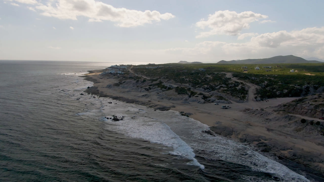 Wide aerial frame over Shipwrecks Beach in Los Cabos, showing gray-blue ocean beside pale sand, darker rocks near the waterline, green scrub inland, distant buildings, and no visible people.