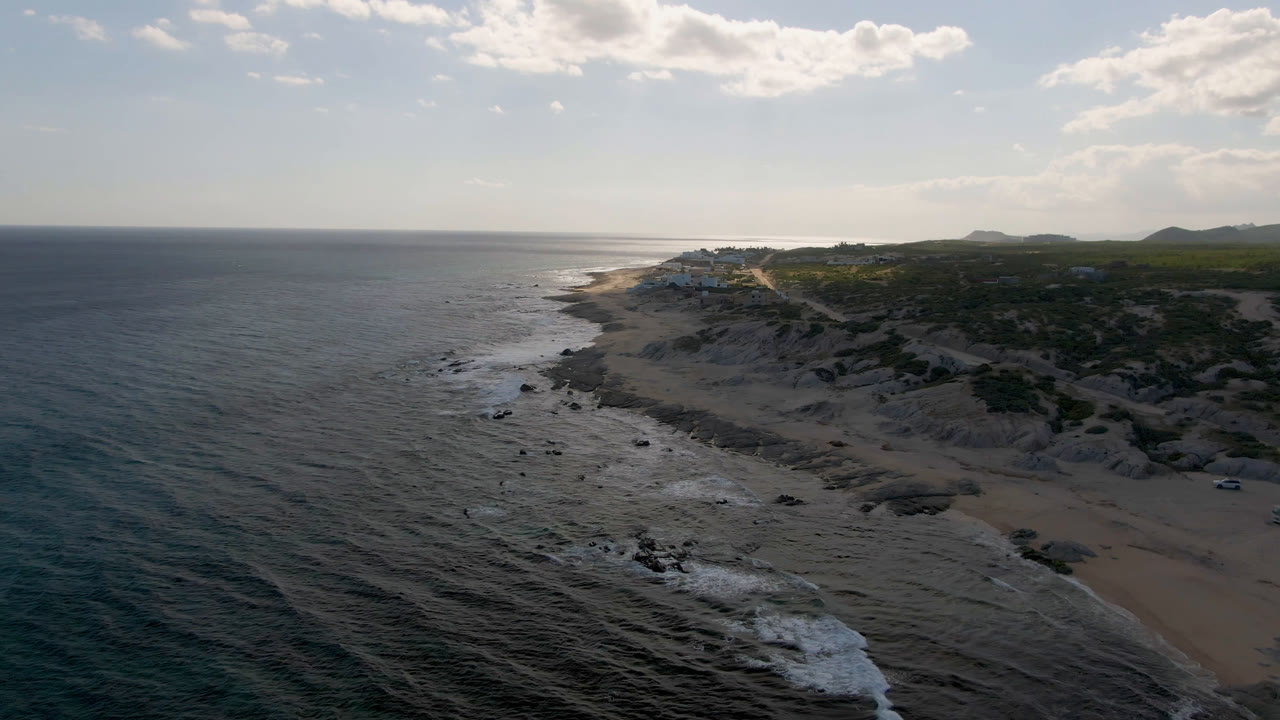 Opening aerial frame over Shipwrecks Beach in Los Cabos, with dark ocean in the foreground, surf curling onto sand and rock, cliffs to the right, and no visible people.