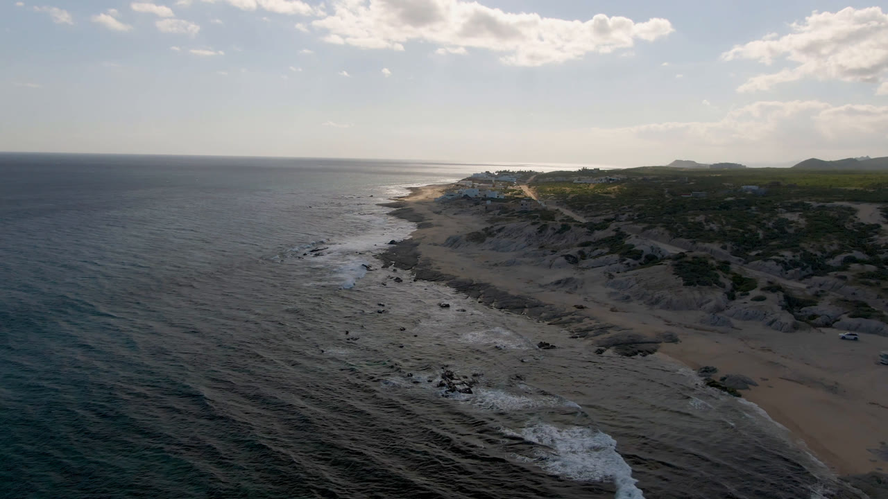 Aerial frame at Shipwrecks Beach in Los Cabos showing more whitewater along the lower beach edge, with the cliff line and scrubland receding toward distant hills and no visible people.