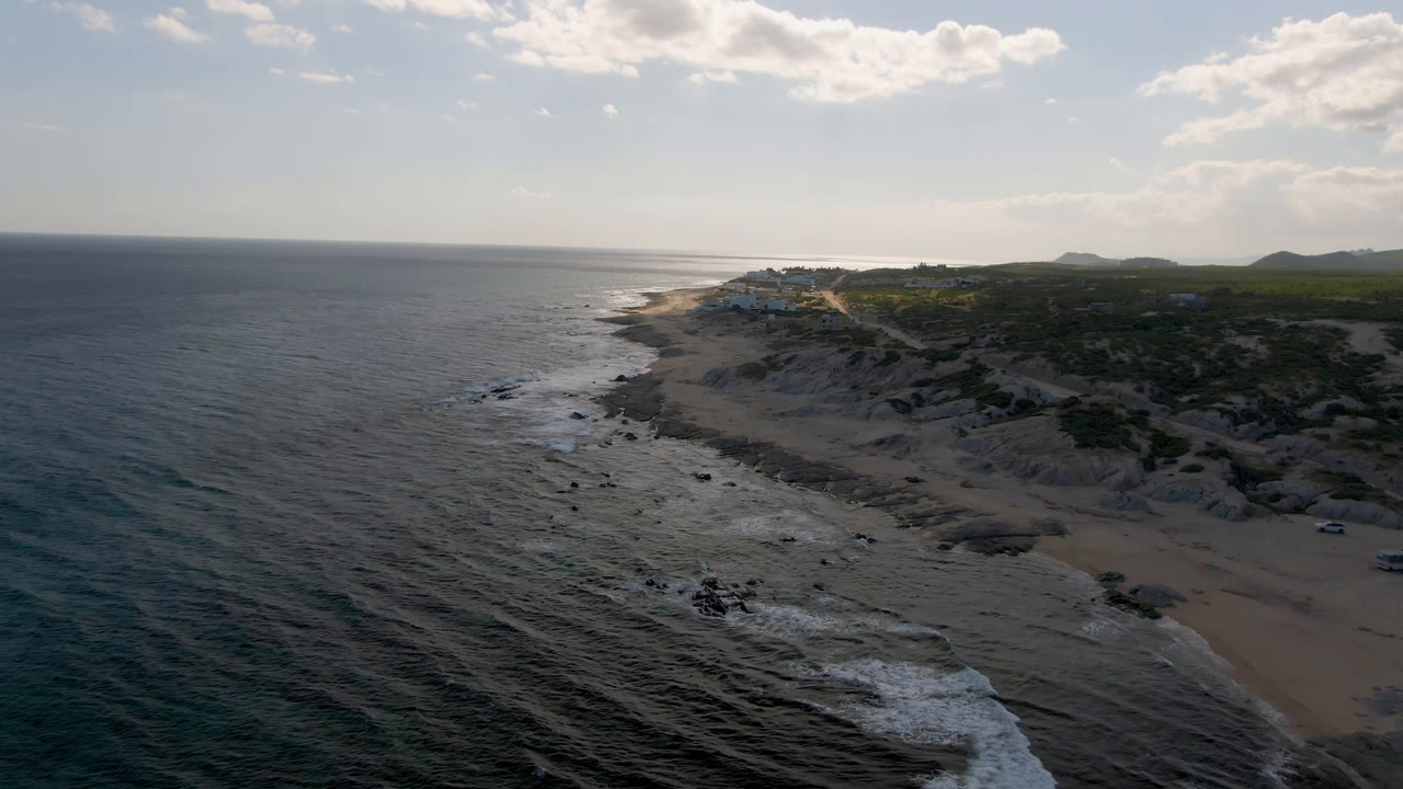 Wide aerial shot of Pacific water beside a rocky sandy shoreline.