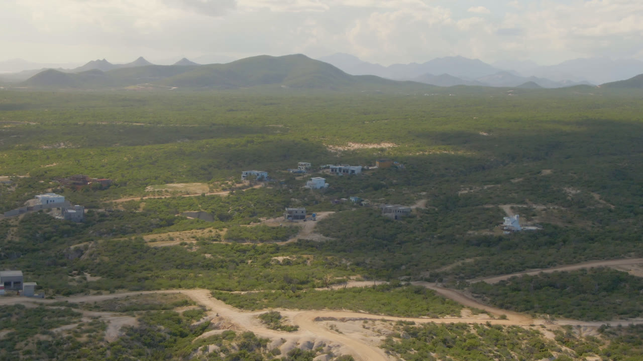 Opening aerial frame over Shipwrecks Beach Los Cabos, Baja California Sur, showing green scrubland, pale dirt roads, scattered buildings, distant mountains, and no visible people.