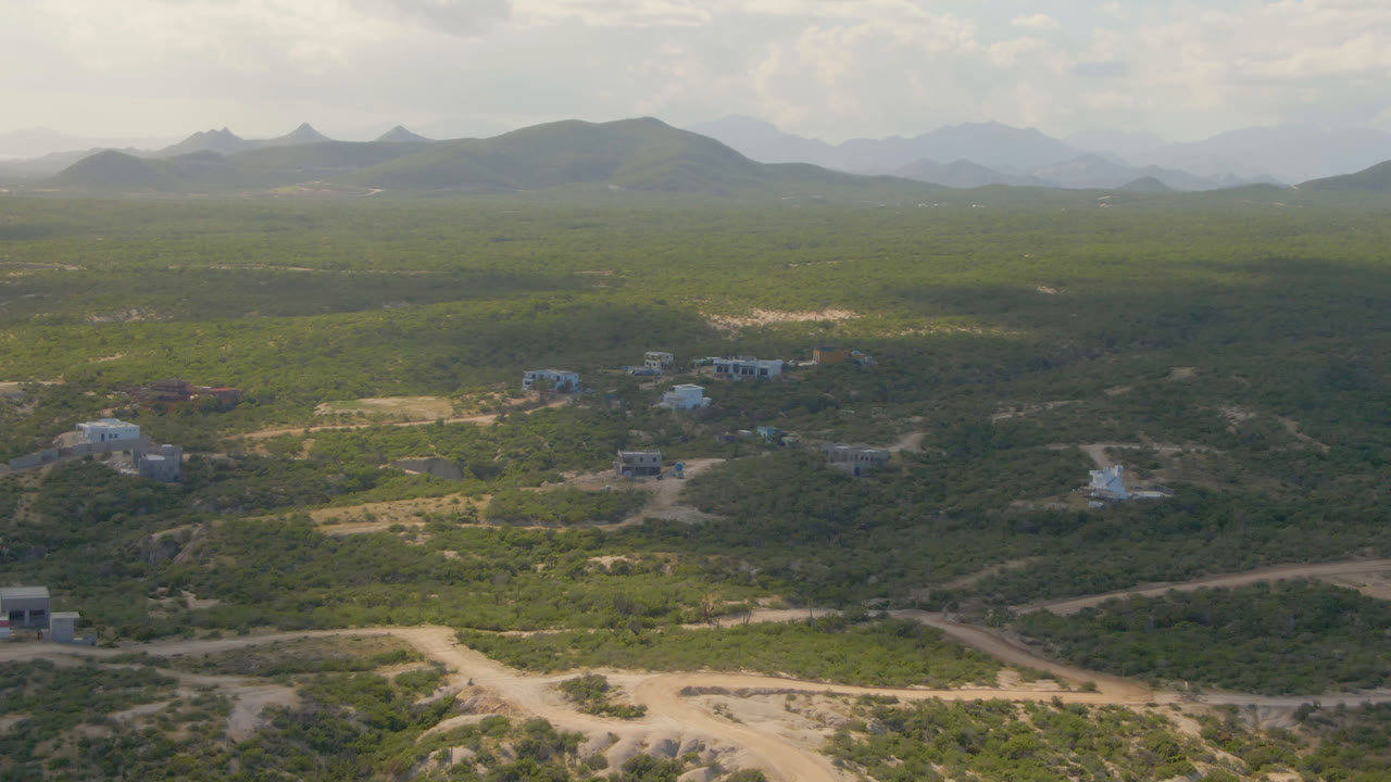Second aerial frame at Shipwrecks Beach Los Cabos with crossing dirt roads in the foreground, small structures in the midground, hazy mountain ridges, and no visible people.