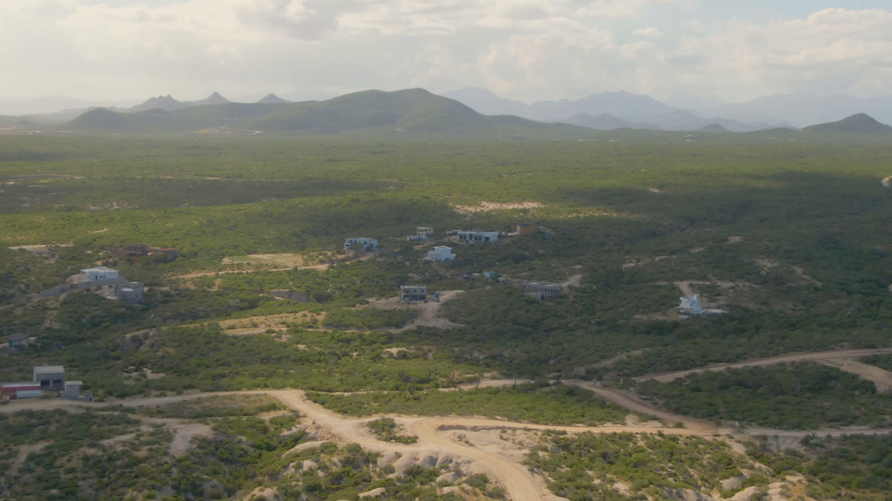 Drone-height view of dirt tracks, low buildings, green terrain, and layered hills.