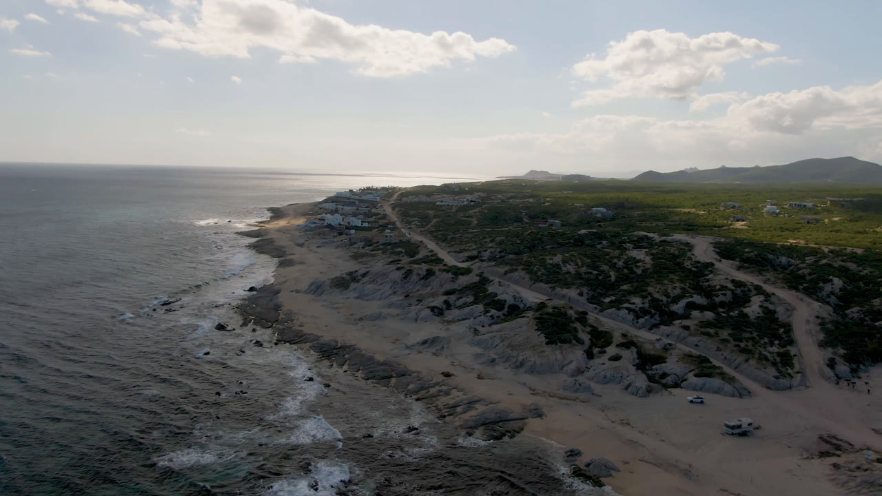 Aerial view of rocky beach, ocean, dirt road, scrubland, and distant hills.
