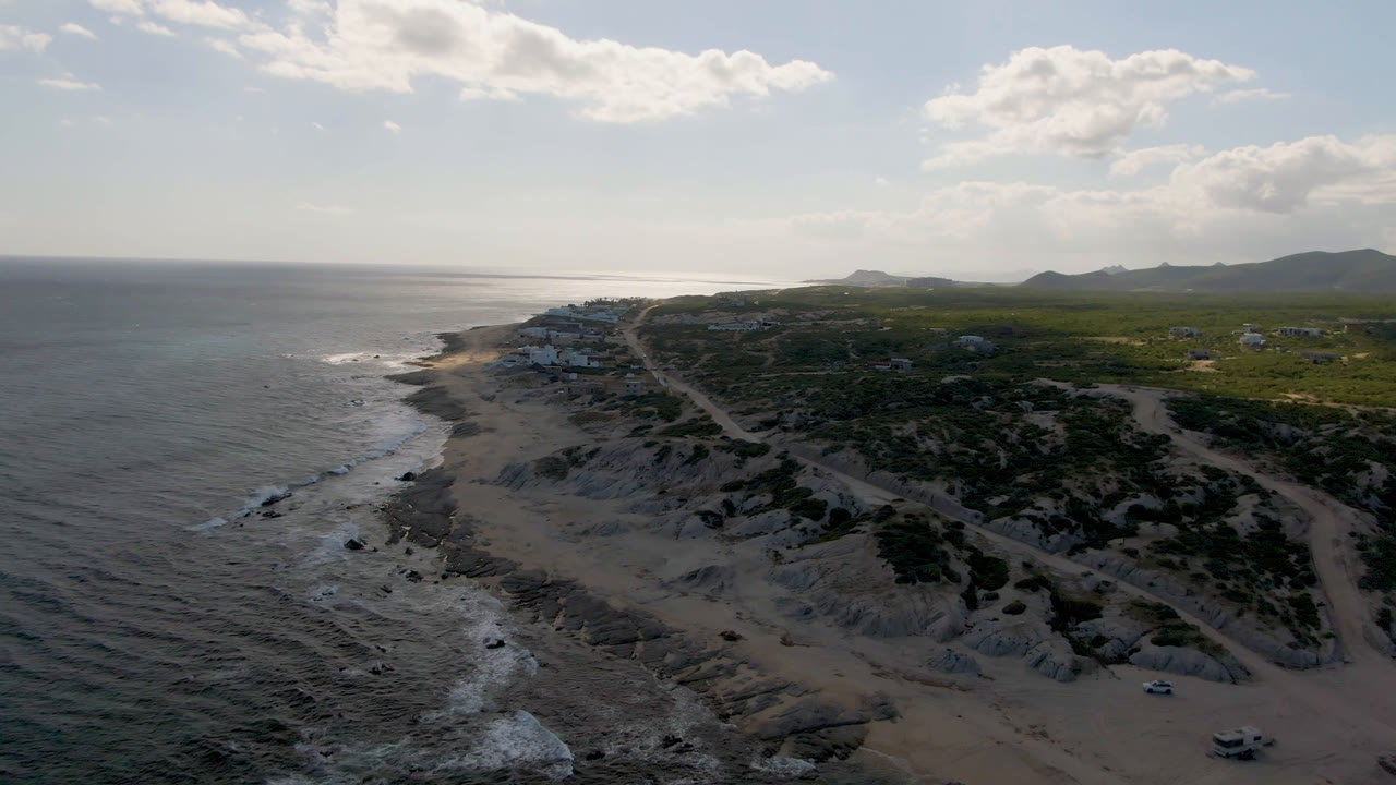 Aerial view along Shipwrecks Beach in Los Cabos showing small waves near the shore, a dirt road running inland from the beach, sparse buildings, green scrub, distant hills, and no visible people.