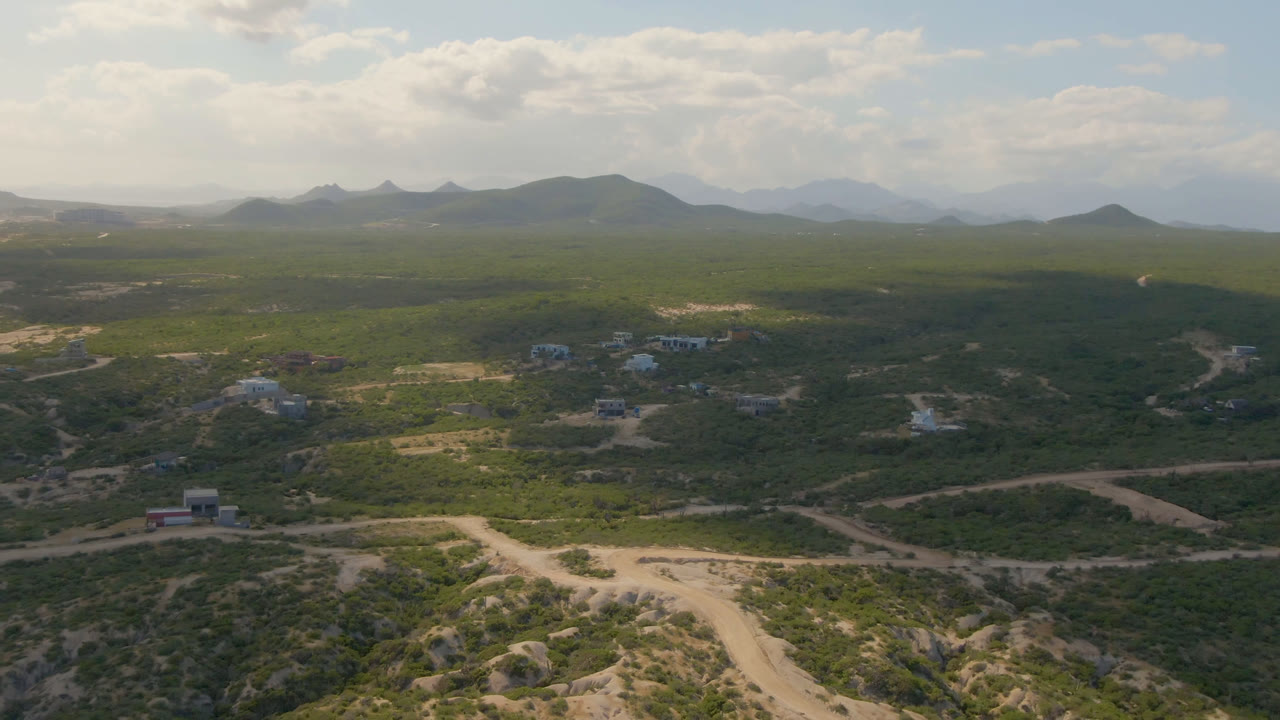 Aerial view over scrub hills, dirt roads, scattered buildings, distant mountains, and cloudy sky.