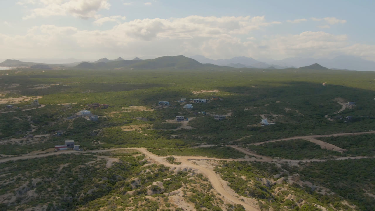 Second aerial frame over Shipwrecks Beach Los Cabos with intersecting dirt roads in the foreground, small buildings in the midground, mountain ridges on the horizon, and no visible people.