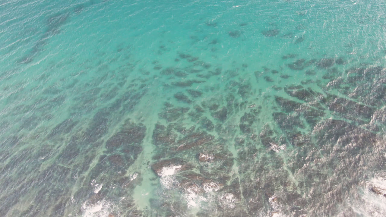 Overhead view of turquoise water and dark reef texture at Shipwrecks Beach in Los Cabos, with white surf near the lower edge and no visible people.