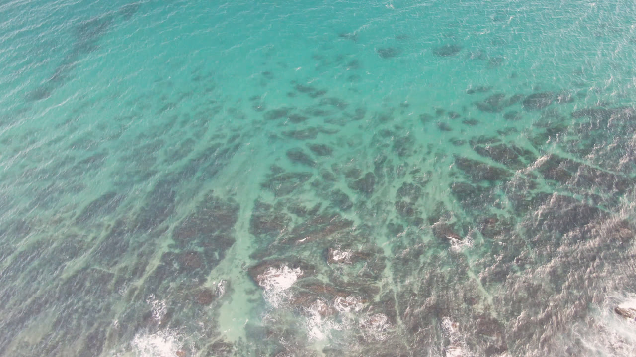 Aerial frame over Shipwrecks Beach water showing blue-green shallows, branching dark reef patches, and small white breakers with no visible people.