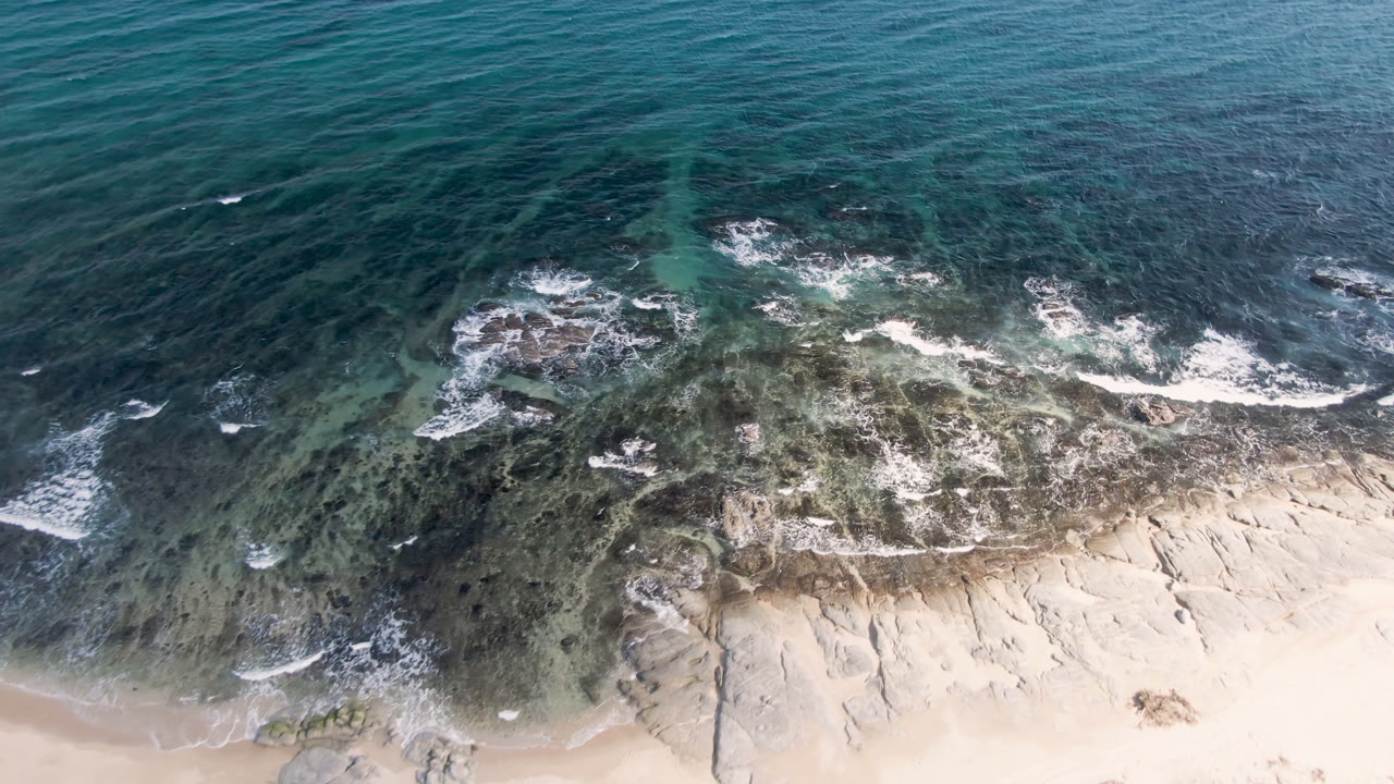 Overhead frame of Shipwrecks Beach in Los Cabos showing blue ocean, white surf over dark reef, pale sand at the bottom edge, and no visible people.