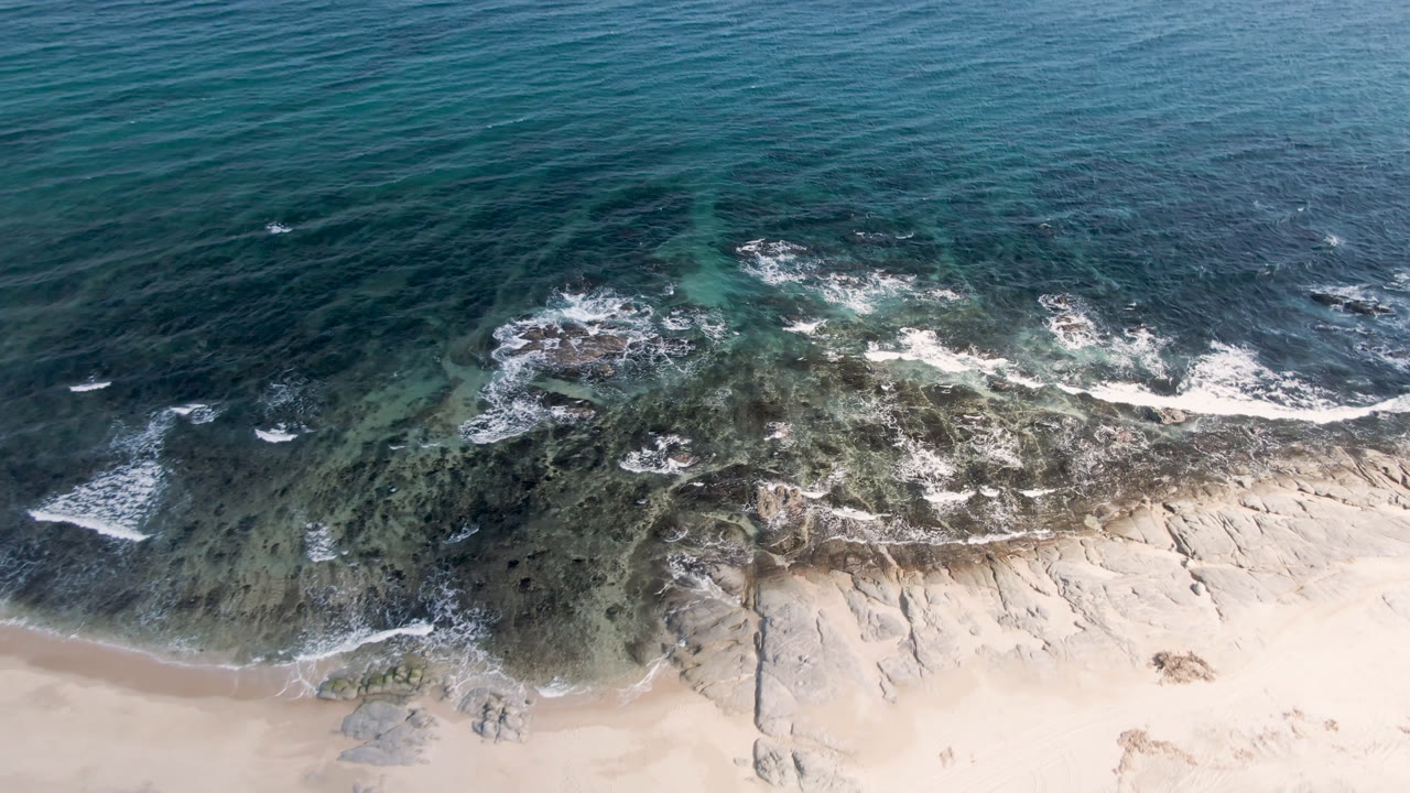 Aerial frame of Shipwrecks Beach in Los Cabos with the surf line spread across the center, deeper blue water above, sand below, and no visible people.