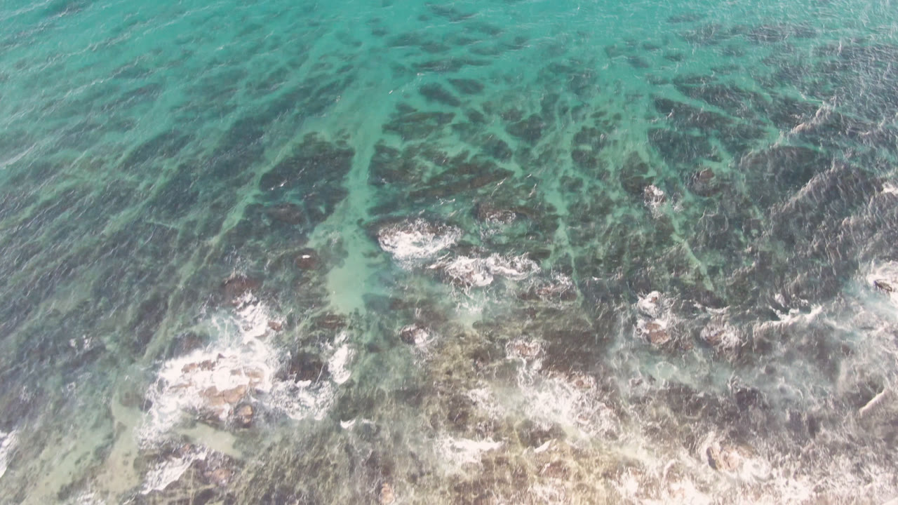 Top-down ocean water breaking over rocks near pale sand.