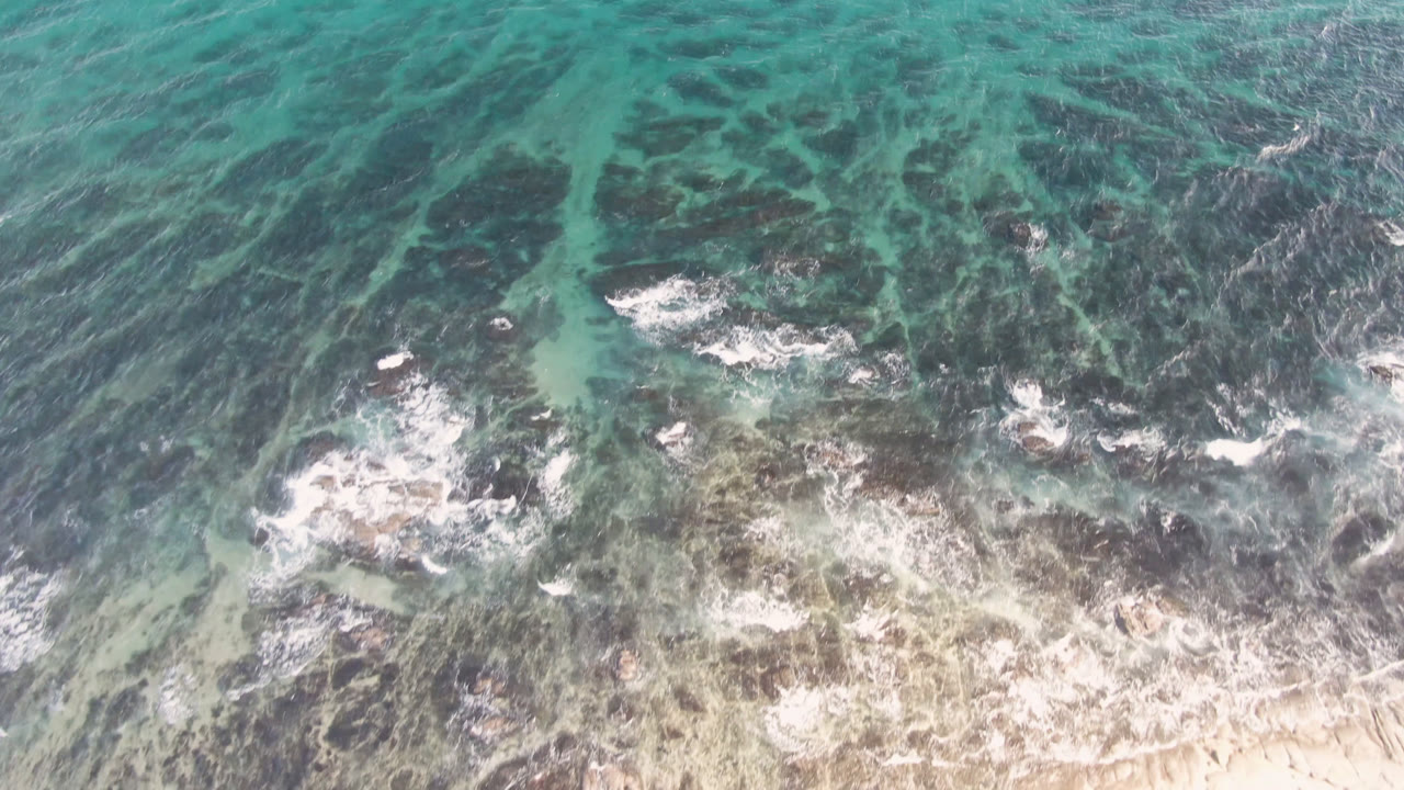 Aerial view over Shipwrecks Beach in Los Cabos with white foam spreading across dark reef patches, green-blue water above, sand along the lower edge, and no visible people.