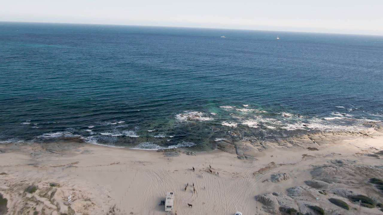 Aerial view at Shipwrecks Beach in Los Cabos with the horizon high in frame, small waves over reef, and pale sand dunes below.