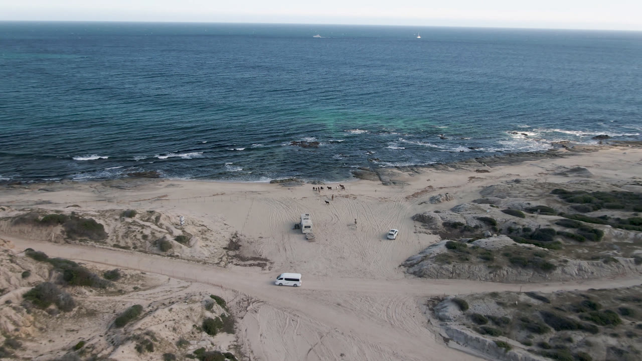 Opening aerial frame over Shipwrecks Beach Los Cabos, with pale sand, scattered rocks, parked white vehicles, blue ocean, and no visible people.