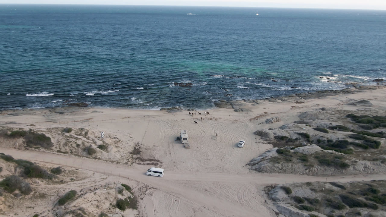Aerial frame at Shipwrecks Beach Los Cabos showing the rocky sand flats nearer center frame, vehicles below the surf line, and no visible people.