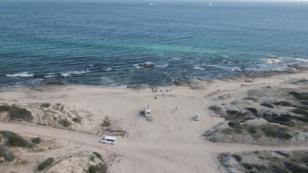 Wide aerial beach view with surf, rocks, sand tracks, and vehicles.