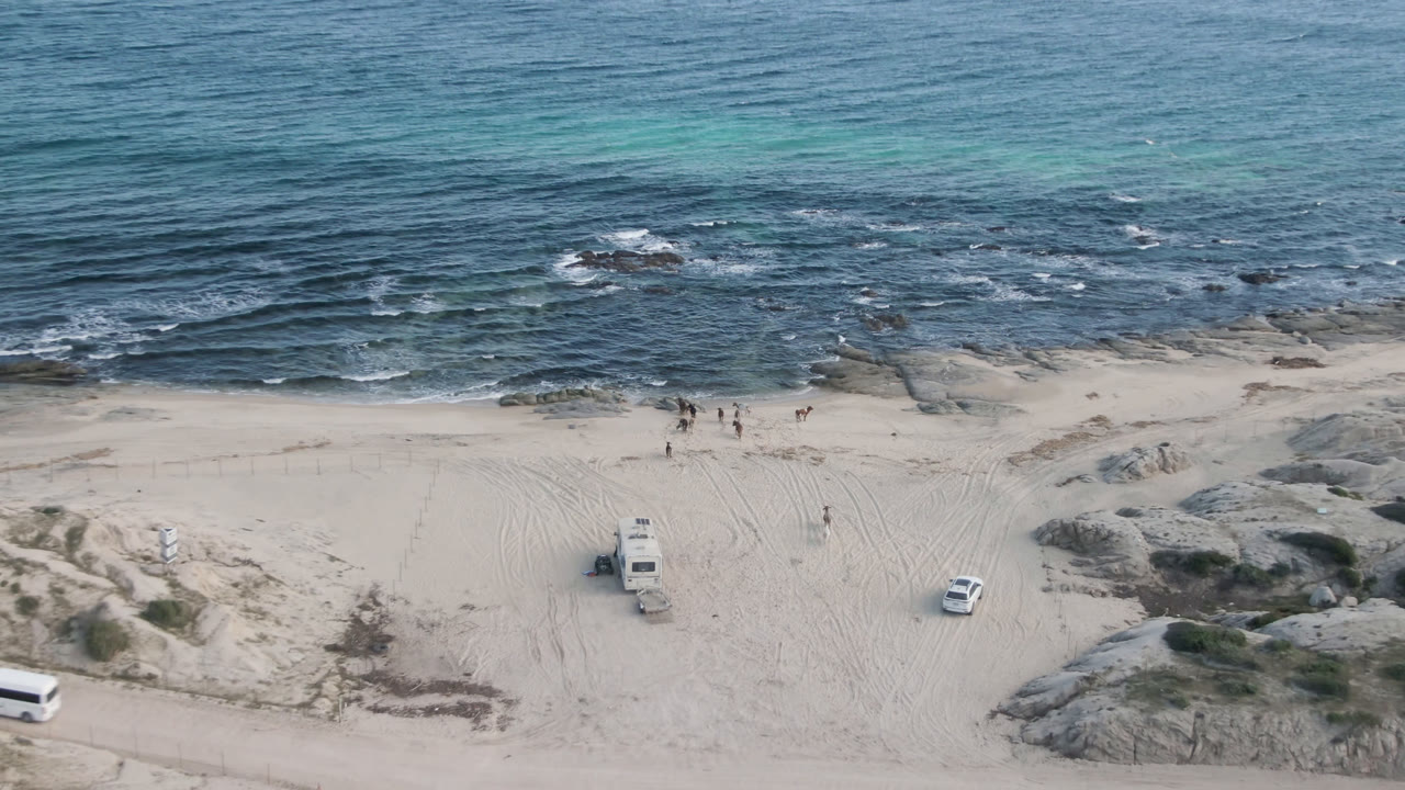 Aerial view of sandy beach, surf, rocks, and two parked vehicles.