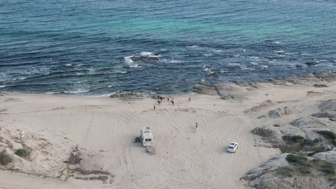 Aerial view at Shipwrecks Beach in Los Cabos with a white vehicle and a boxy beach vehicle on marked sand, rock shelves at the edge of the surf, and small distant figures by the water.