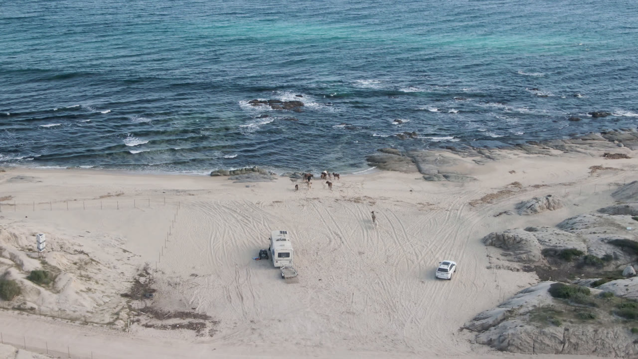 Aerial frame at Shipwrecks Beach in Los Cabos showing blue waves above pale sand, rocky outcrops, dunes, tire tracks, a white vehicle, and a larger beach vehicle with no visible people.