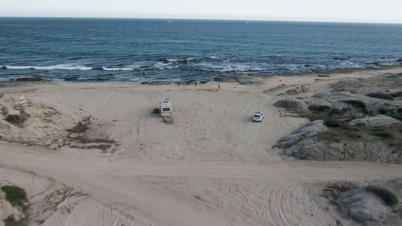 Opening aerial frame at Shipwrecks Beach in Los Cabos shows a white RV left of center, a white car to the right, pale sand, rocky shoreline, blue surf, and no visible people.