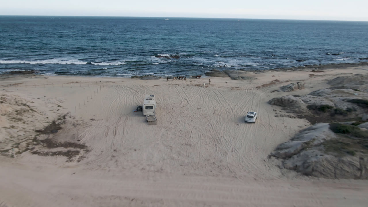 Second frame keeps the white RV and car separated on the sand at Shipwrecks Beach, with tire marks, low dune mounds, dark rock shelves, ocean waves, and no visible people.