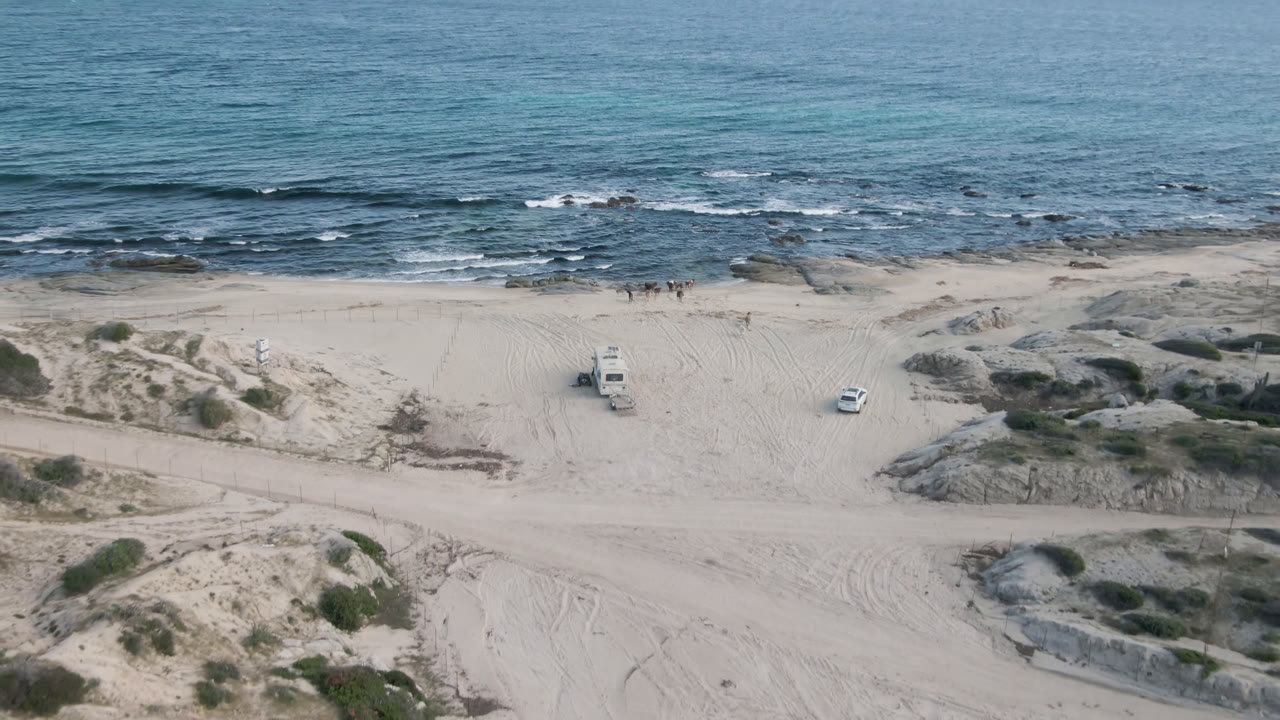 At Shipwrecks Beach in Los Cabos, a light-colored truck is parked on pale sand below low dunes, with rocky surf and blue ocean behind it and no visible people.