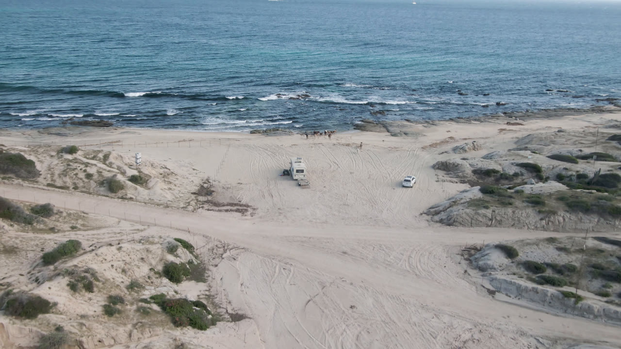 The Shipwrecks Beach shoreline remains wide in frame, with the parked truck near center-left, tire-marked sand in the foreground, dark rocks at the waterline, and no visible people.