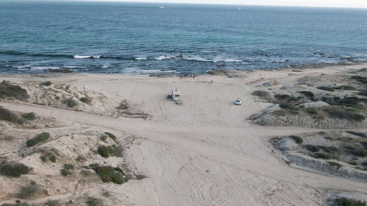 Aerial beach view with truck, dunes, tire tracks, and ocean horizon.