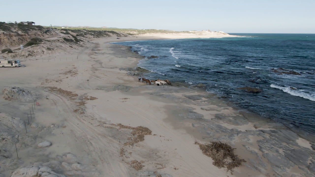 Shipwrecks Beach in Los Cabos seen from above, with the small boat still near the rocky shoreline, thin waves moving across the sand, distant dunes, and no visible people.