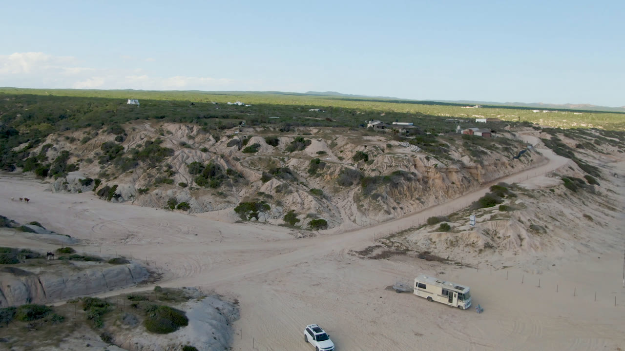 Opening aerial frame over Shipwrecks Beach in Los Cabos, Mexico, with a camper truck near sandy tracks, scrub-covered dunes, distant camps, and no visible people.