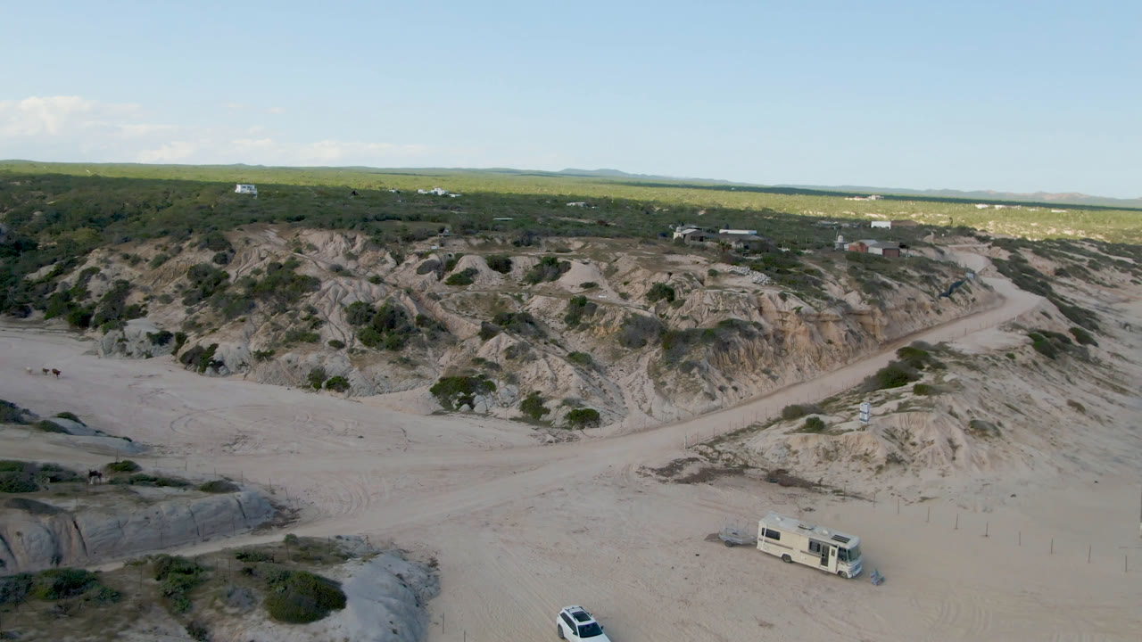 A wide aerial view at Shipwrecks Beach in Los Cabos, Mexico, showing eroded sandy dunes, a camper truck lower in frame, distant structures, and no visible people.