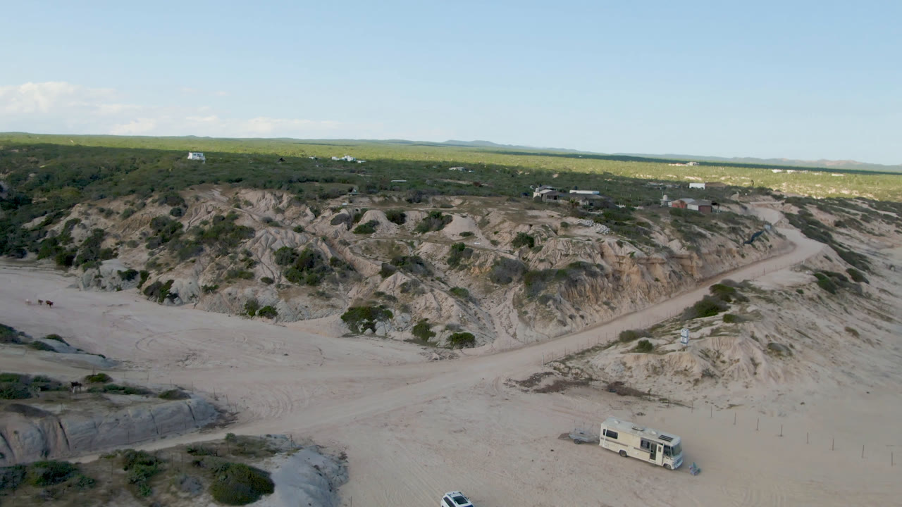 Aerial coastal dune landscape with camper truck, sandy road, and low buildings.