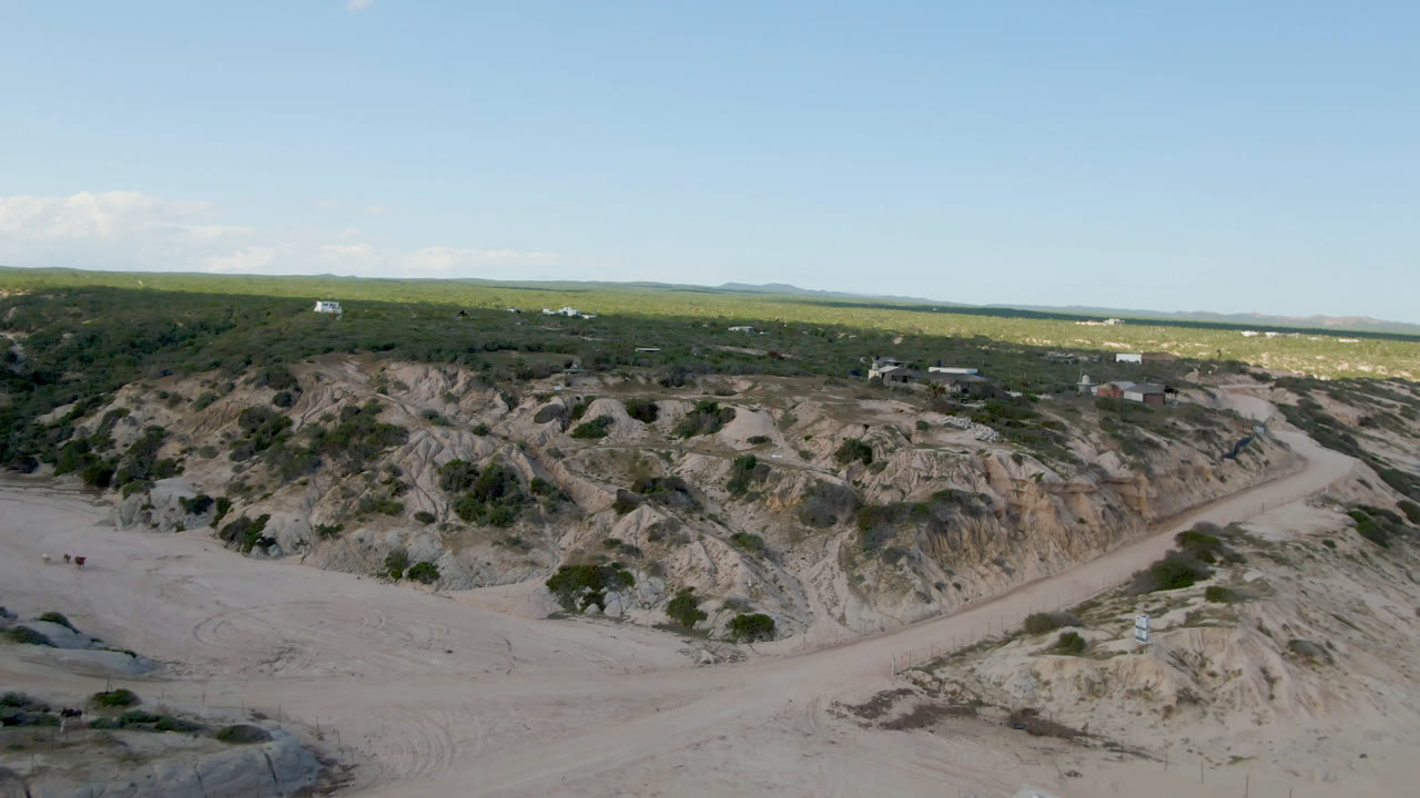 Aerial frame at Shipwrecks Beach Los Cabos showing a pale dirt road beside eroded rock and scrub, with ocean on the horizon and no visible people.