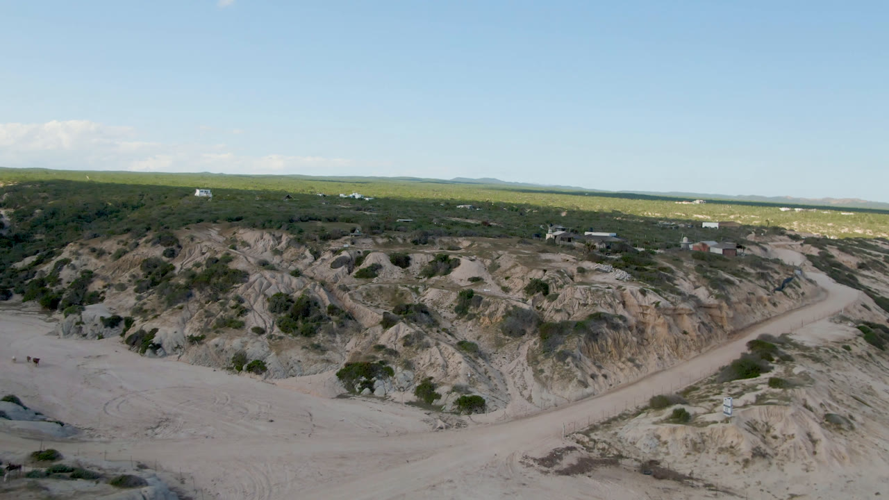 Aerial frame at Shipwrecks Beach Los Cabos with the winding dirt track more centered between pale rock cuts, scrub patches, distant water, and no visible people.