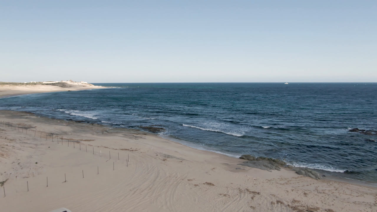 Opening frame of Shipwrecks Beach Los Cabos with pale sand, fence posts, blue ocean, low dunes at left, a distant boat on the horizon, and no visible people.