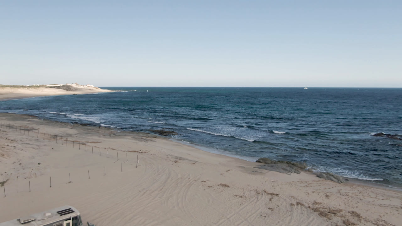 Second frame at Shipwrecks Beach Los Cabos showing sand tracks, fence posts, surf lines, a partial camper near the lower edge, and no visible people.