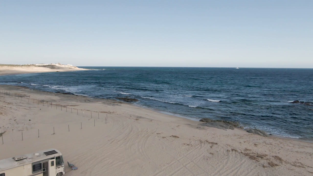 Aerial beach view with surf, rocks, fence posts, and a camper corner.