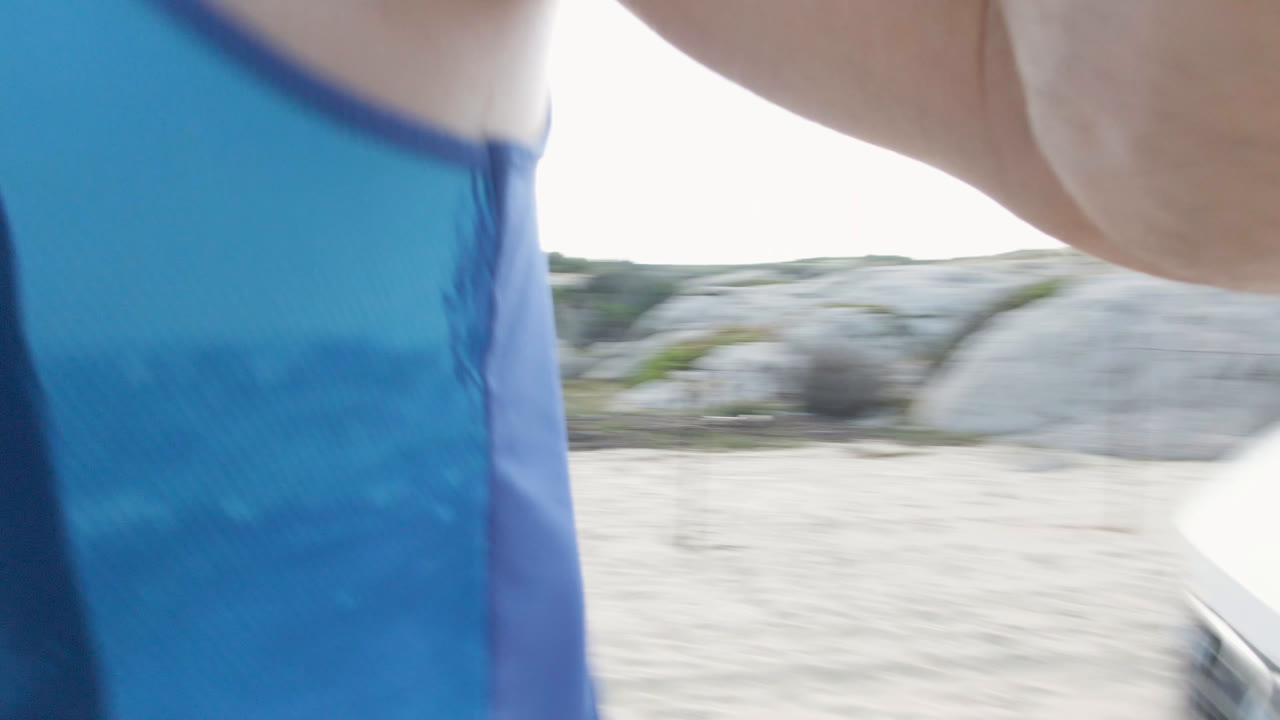 At Shipwrecks Beach in Los Cabos, a partly visible person in blue and white clothing stands close to a white vehicle on sand, with bright sky and beach glare behind them.