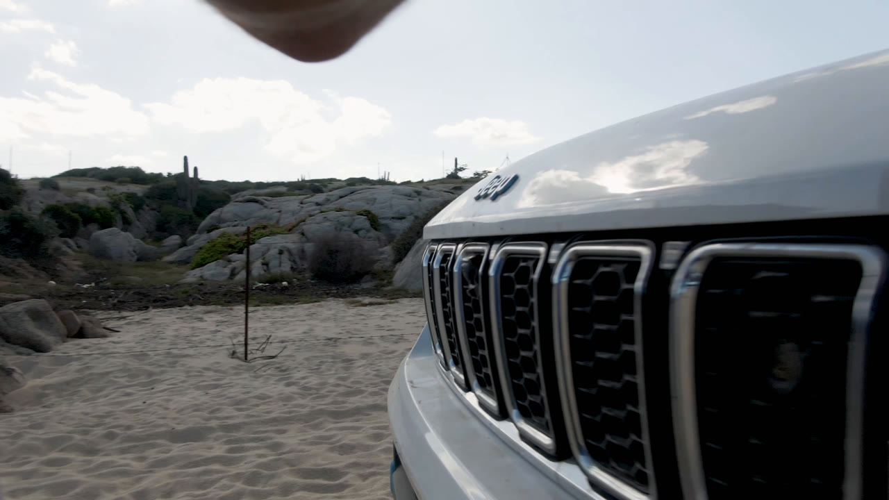 At Shipwrecks Beach in Los Cabos, the front grille of a white Jeep sits close to the camera, with sandy ground, pale rocks, low vegetation, and bright daylight in the background.