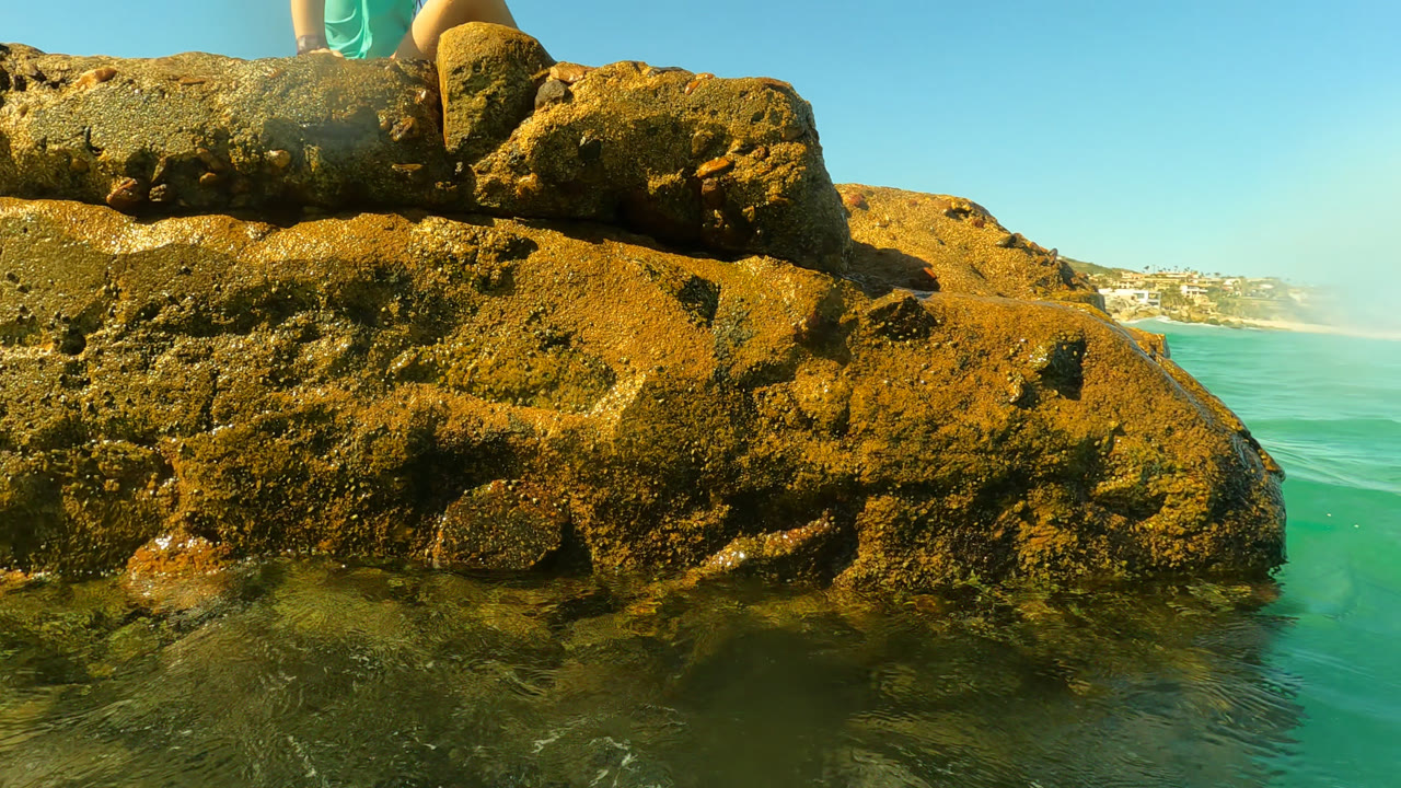 A low view in Palmilla, Baja California Sur shows barnacled-looking rocks and shallow turquoise water, with the lower legs of a seated person cropped at the top edge.