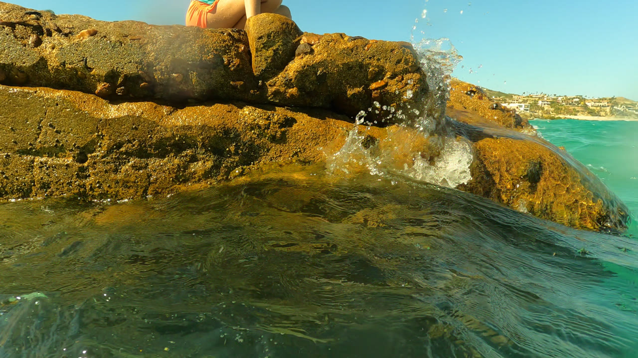 At Palmilla, Baja California Sur, a water-level view shows green-blue ocean pushing toward ochre rocks while a cropped seated woman is partly visible at the top edge.