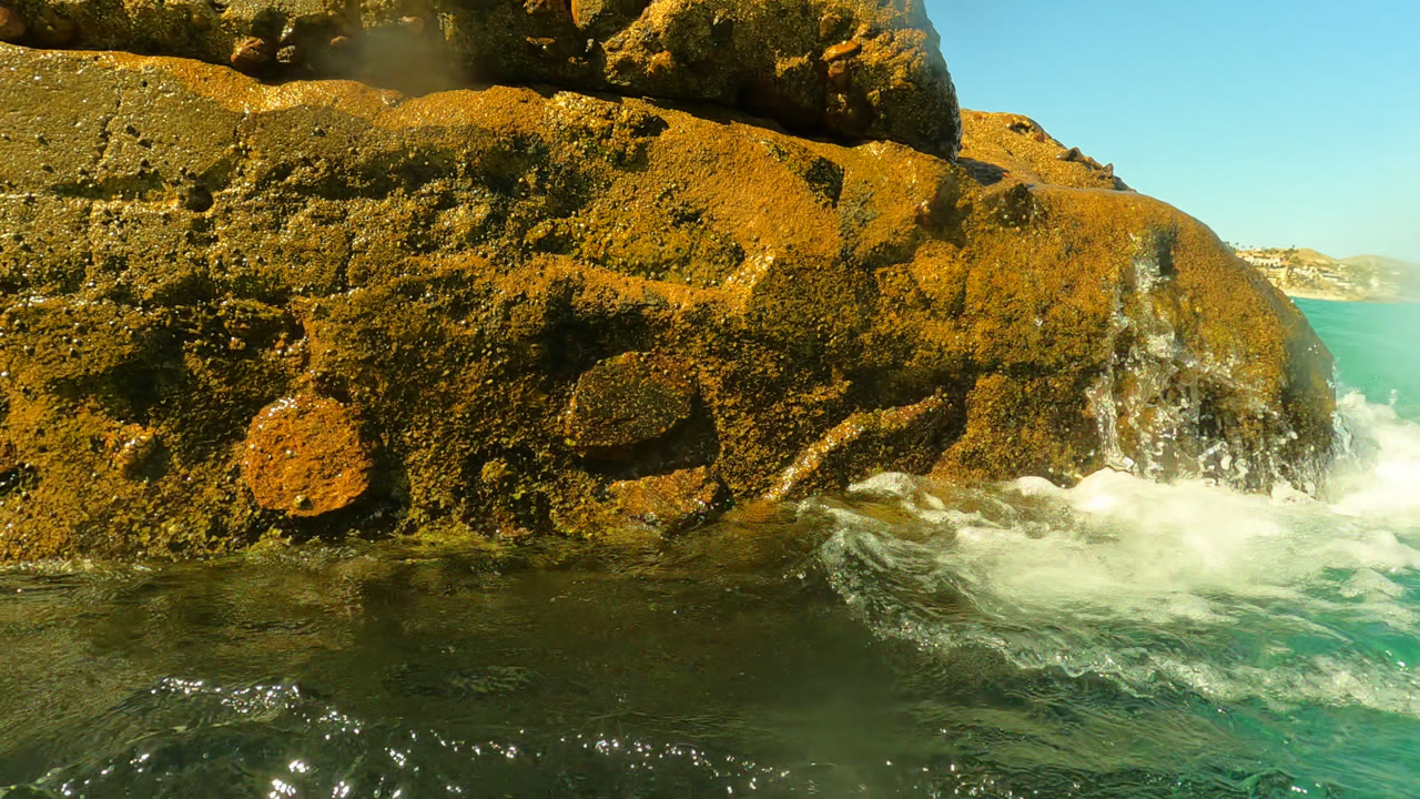 At Palmilla, Baja California Sur, foamy water breaks at the base of a rough ochre rock while the distant coastline and blue sky sit behind it.
