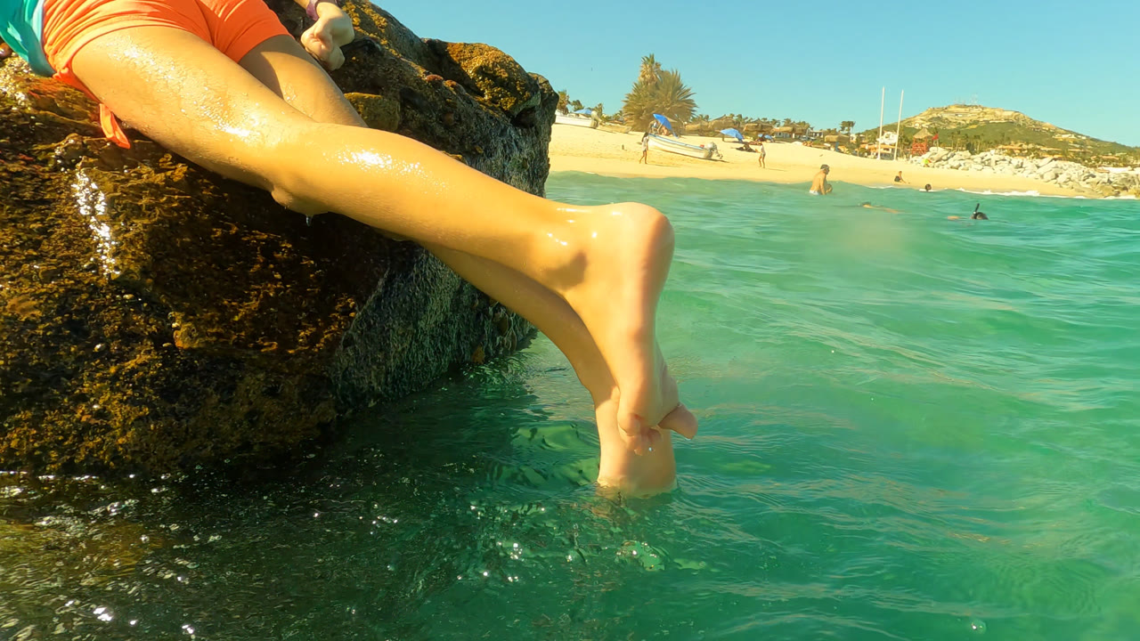 At Palmilla, Baja California Sur, bare legs extend beside dark algae-covered rocks as clear turquoise water fills the foreground and a sandy beach sits in the distance, with no visible faces.